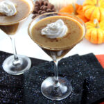 Paleo Pumpkin Spice Martinis - near pine cones, pumpkins, and sparkly black napkin - topped with coconut whipped cream.