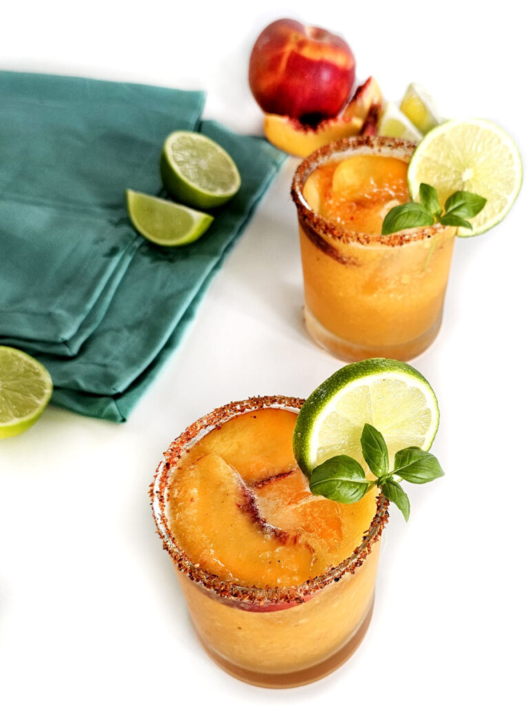 Two Hot Honey Peach Margaritas and peach slices and blue napkin and limes