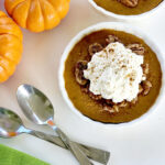 Pumpkin custard with caramelized pecans, whipped cream, two spoons, a green napkin, and mini pumpkins in background
