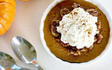 Pumpkin custard with caramelized pecans, whipped cream, two spoons, a green napkin, and mini pumpkins in background