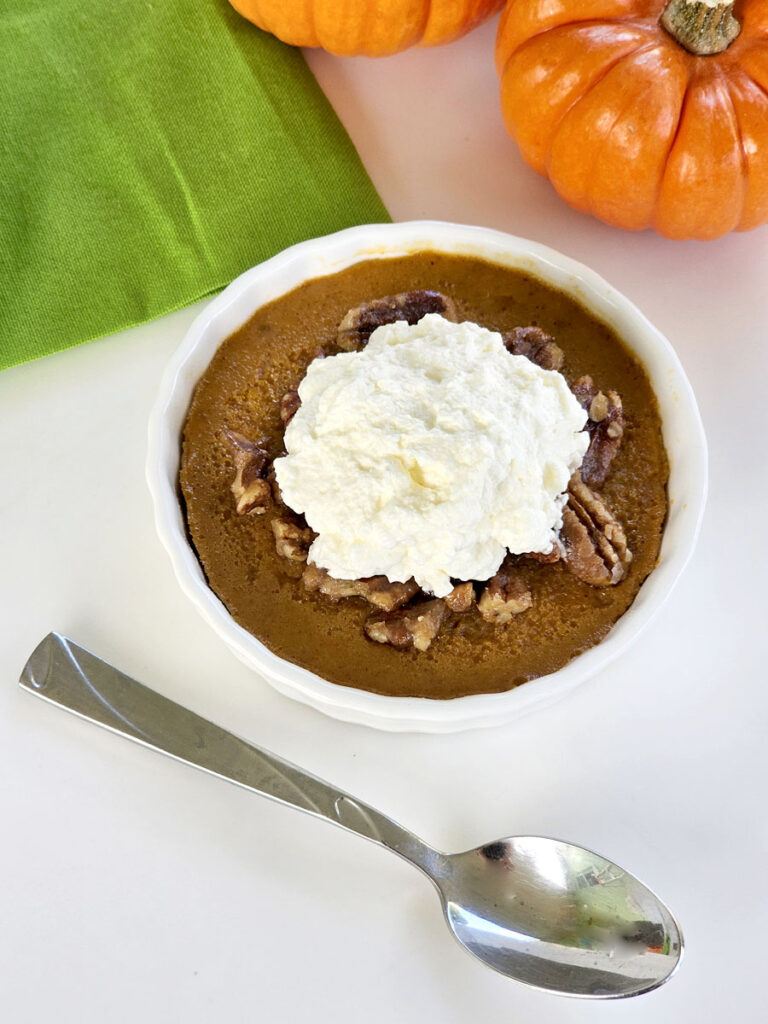 Pumpkin custard with caramelized pecans, whipped cream, a spoon, a green napkin, and mini pumpkins in background