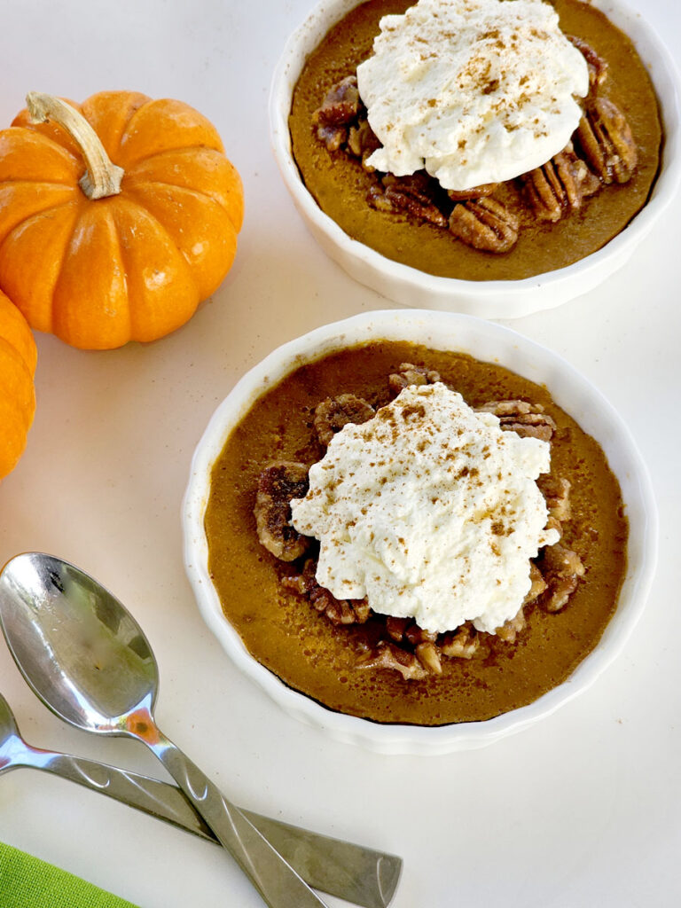 Pumpkin custard with caramelized pecans, whipped cream, two spoons, and mini pumpkins in background