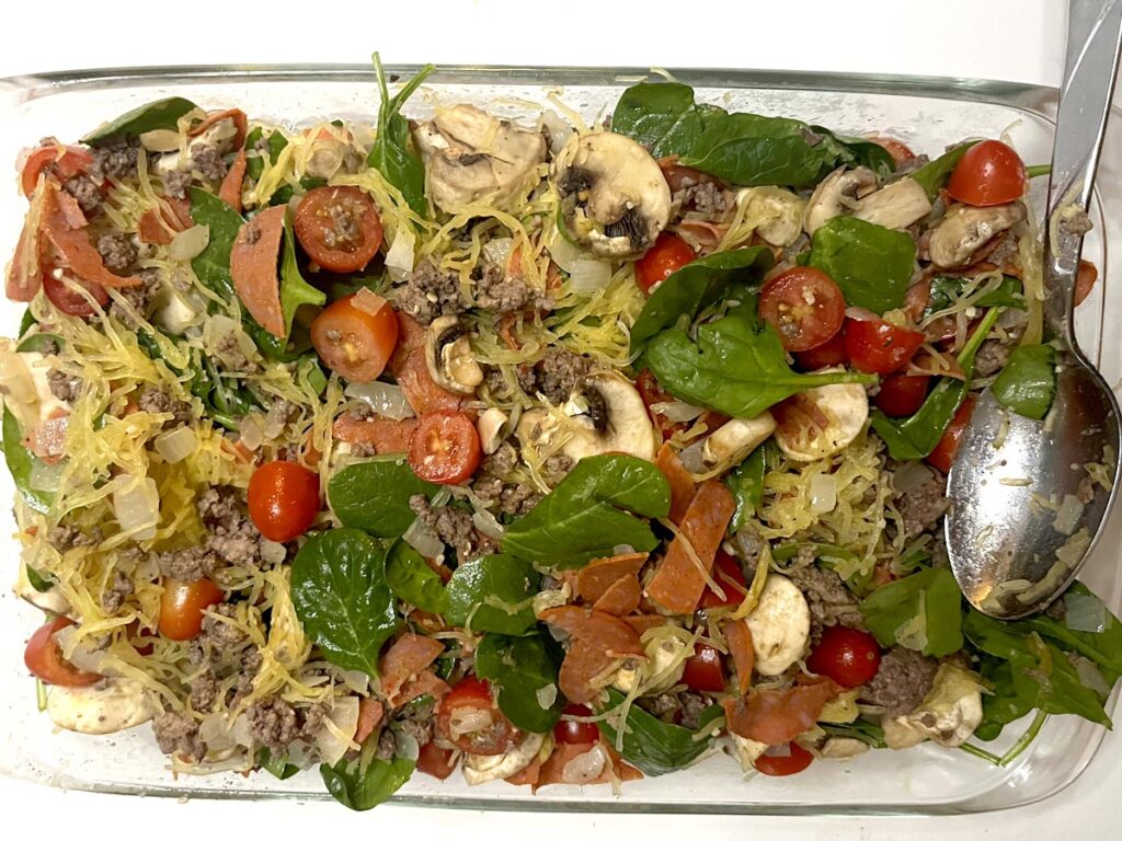 9x13 glass pan with vegetables, meats, and spaghetti squash added with a large silver spoon for stirring