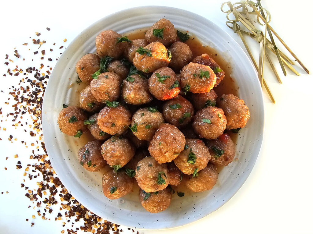 Grape jelly chili meatballs on white platter surrounded by chili flakes and party skewers