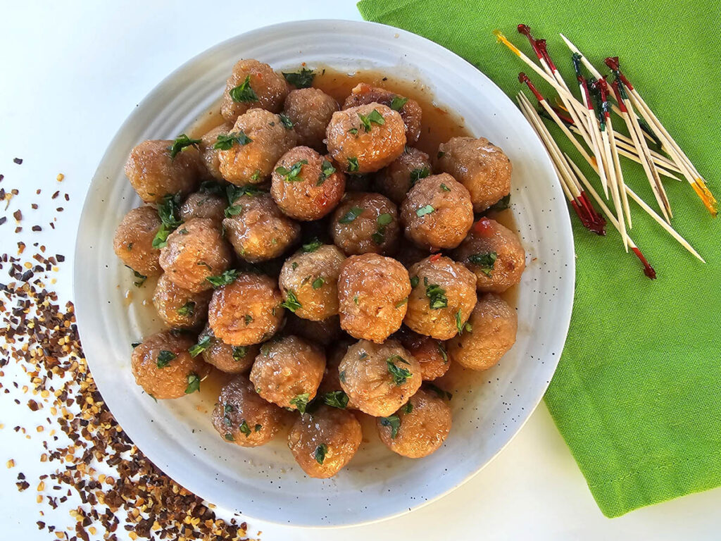 Grape jelly chili meatballs on white platter surrounded by chili flakes and party skewers next to green napkin