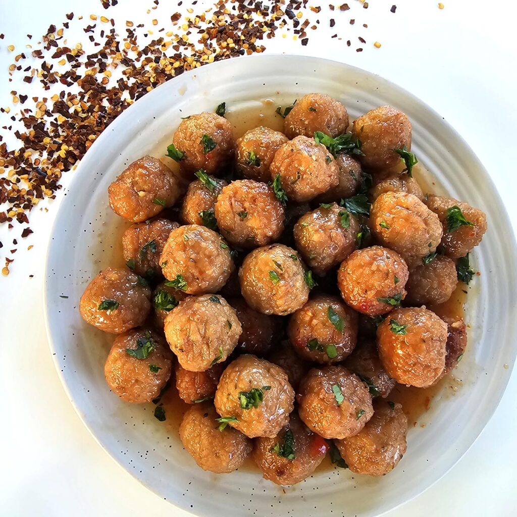 Grape jelly chili meatballs on white platter surrounded by chili flakes and party skewers