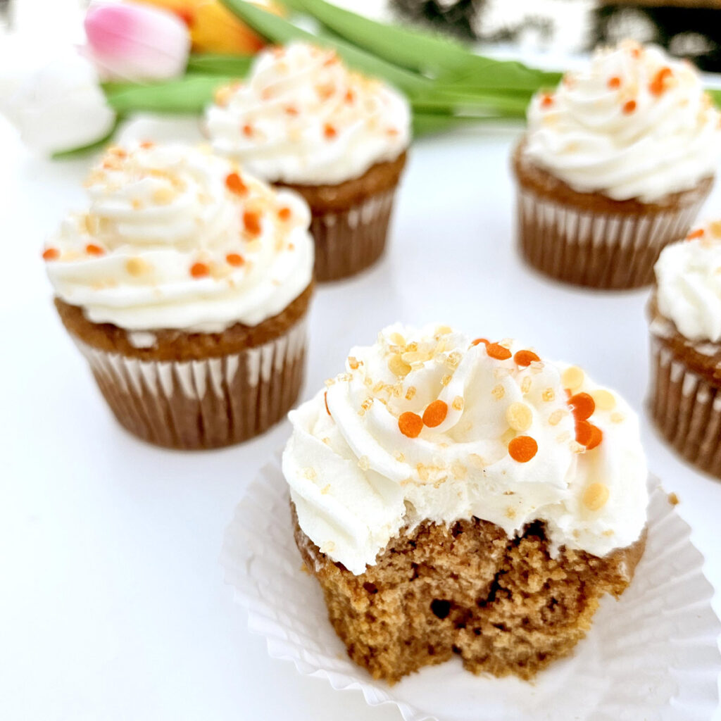 Carrot cake cupcakes with orange sprinkles next to blue napkins and in front of colorful tulips. With bite taken out of one.