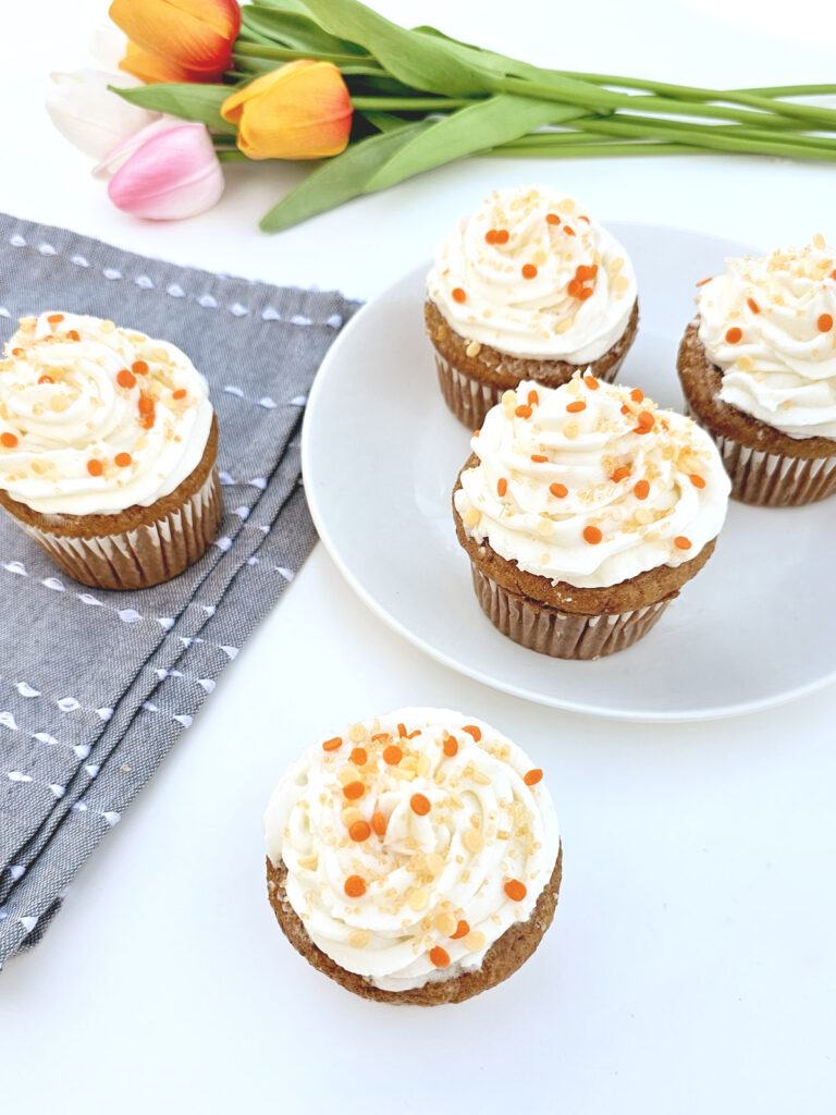 Carrot cake cupcakes with orange sprinkles next to blue napkins and in front of colorful tulips.
