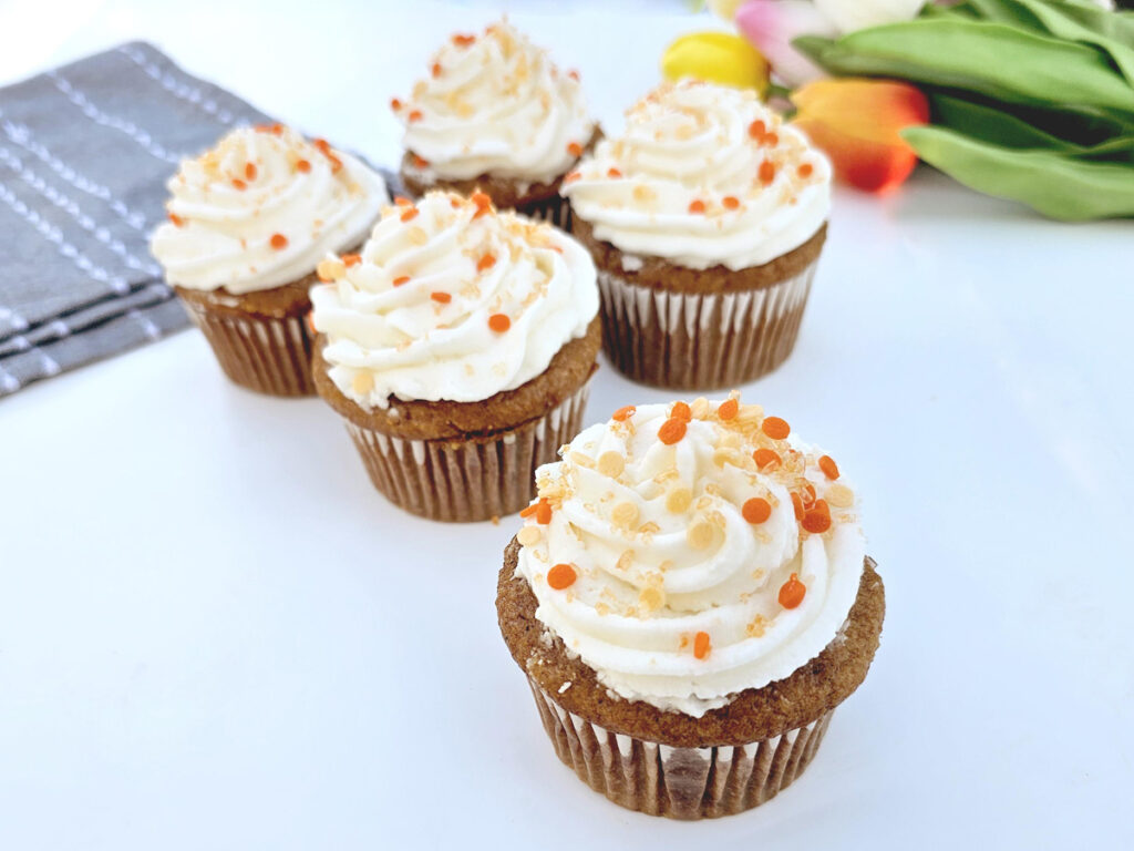 Carrot cake cupcakes with orange sprinkles next to blue napkins and in front of colorful tulips.