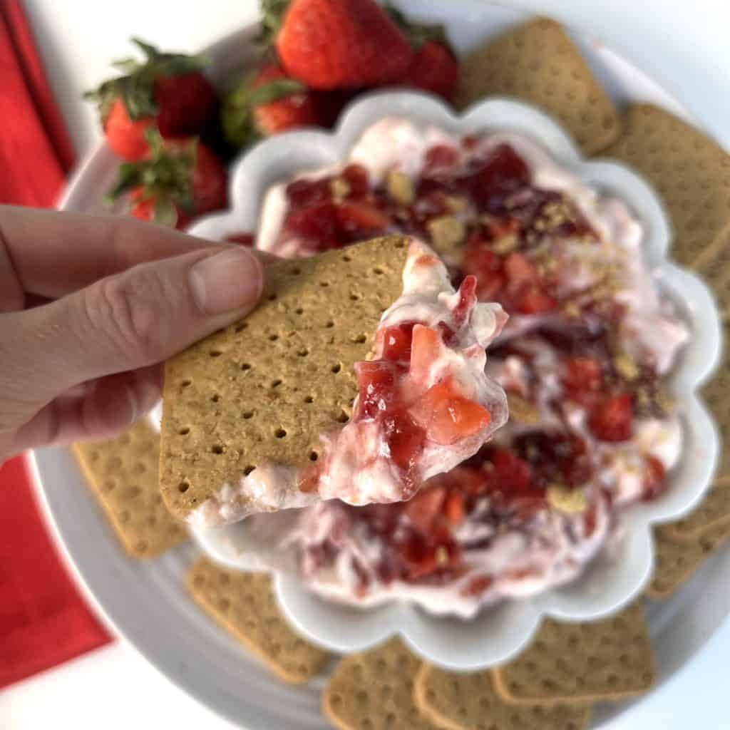 Strawberry Cheesecake Dip served with strawberries and graham crackers on a white plate inside of white flower shaped bowl with white dip, strawberry jam swirls, sprinkle in diced strawberry and graham cracker crumbs. Graham cracker dipping in it.