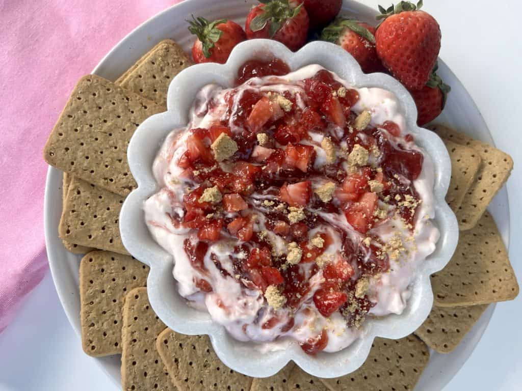 Strawberry Cheesecake Dip served with strawberries and graham crackers on a white plate inside of white flower shaped bowl with white dip, strawberry jam swirls, sprinkle in diced strawberry and graham cracker crumbs.