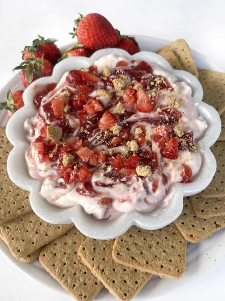 Strawberry Cheesecake Dip served with strawberries and graham crackers on a white plate inside of white flower shaped bowl with white dip, strawberry jam swirls, sprinkle in diced strawberry and graham cracker crumbs.